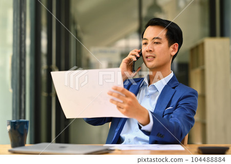 Asian male financial advisor looking at document, consulting client distantly by mobile phone call 104488884