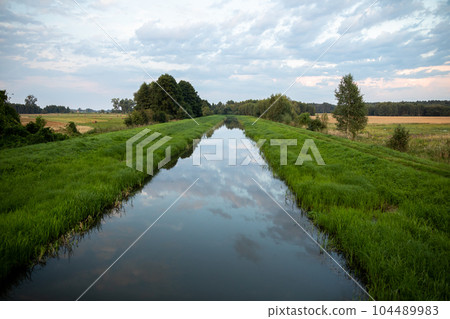 A slow-flowing wide river among lush green grasses and agricultural fields. 104489983
