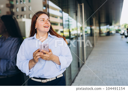 Businesswoman using phone near office. Happy woman in a blue shirt walks on the street with her smartphone. Professional manager working on mobile device. Successful female using modern technology Businesswoman using phone near office. Happy woman in a blue shirt walks on the street with her smartphone. Professional manager working on mobile device. Successful female using modern technology 104491154