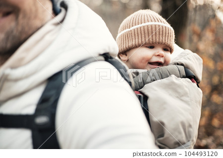 Sporty father carrying his infant son wearing winter jumpsuit and cap in backpack carrier hiking in autumn forest 104493120