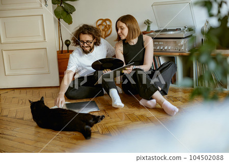 Couple sitting on floor and stroking cats while listening music on vinyl player with records 104502088
