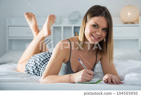 Young smiling girl lounging in bed and writing to notebook Young smiling girl lounging in bed and writing to notebook 104503950