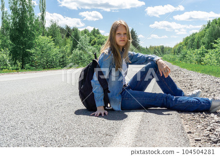 A young girl with a backpack is sitting on the side of the road waiting for vehicles. The concept of travel, hitchhiking, stupidity, vacation. A young girl with a backpack is sitting on the side of the road waiting for vehicles. The concept of travel, hitchhiking, stupidity, vacation. 104504281