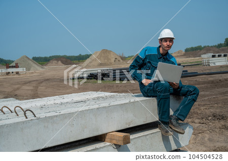 Caucasian male builder in hardhat sits on floor slabs and uses laptop at construction site.  104504528