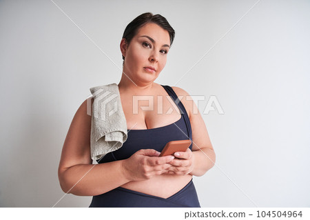 Sporty fat woman holding smartphone and poses camera in white wall 104504964