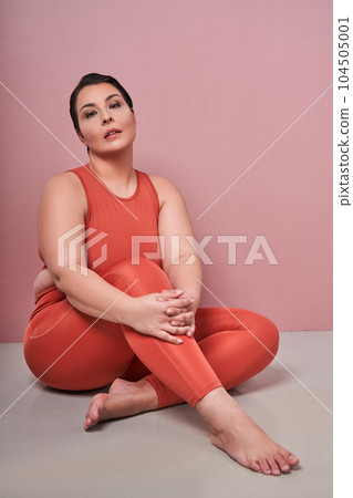 Confident woman in sportswear looking at camera and sitting over pink studio wall 104505001