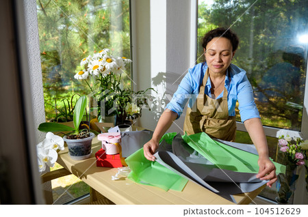 Florist at work. Charming multi ethnic adult woman in apron, rolling out wrapping paper on the table while creating a beautiful bouquet of flowers, for festive life event in a floral design studio 104512629