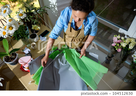 Top view of a pregnant woman, inspired florist entrepreneur in apron, rolling out wrapping paper on the table while creating a beautiful bouquet of flowers, for festive life event in a floral store Top view of a pregnant woman, inspired florist entrepreneur in apron, rolling out wrapping paper on the table while creating a beautiful bouquet of flowers, for festive life event in a floral store 104512630