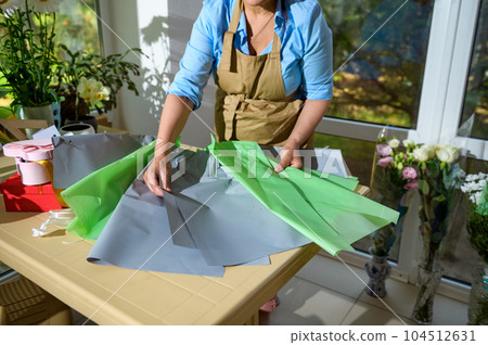 Close-up of florist laying color wrapping paper on the table while arranging flower bouquet in floral design studio. Small business. Flower shop. Making decorations and arrangements. Flowers delivery Close-up of florist laying color wrapping paper on the table while arranging flower bouquet in floral design studio. Small business. Flower shop. Making decorations and arrangements. Flowers delivery 104512631