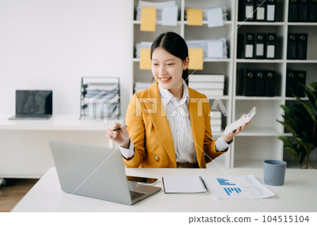 Young attractive Asian woman smiling thinking planning writing in notebook, tablet and laptop working from home, looking at camera at office 104515104