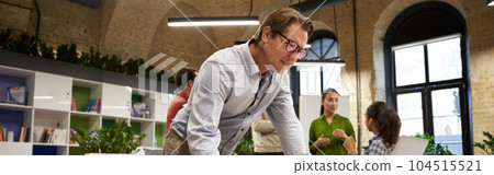 Successful businessman standing in the modern coworking space, looking at laptop screen and thinking about something while his young colleagues talking on the background 104515521