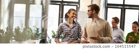 Two young diverse business people, male and female coworkers talking, discussing project and smiling while walking down the modern office Two young diverse business people, male and female coworkers talking, discussing project and smiling while walking down the modern office 104515547