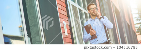 Business talk. Young confident caucasian businessman wearing blue shirt and eyeglasses holding his laptop and talking by phone while walking outdoors 104516783