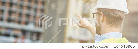 Building industry. Rear view of a young civil engineer or construction supervisor wearing helmet pointing at construction site Building industry. Rear view of a young civil engineer or construction supervisor wearing helmet pointing at construction site 104516809