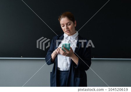 Caucasian female teacher in a suit uses a smartphone while standing at the blackboard. 104520619
