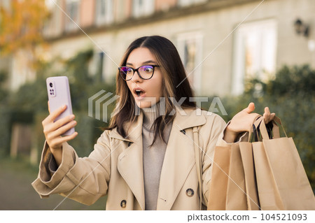 Shocking sales. Surprised fashionable woman checking her phone holding paper shopping bags in the street 104521093
