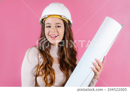 Portrait of a happy female architect builder in the hard hat holding layout plan on the pink background. Portrait of a happy female architect builder in the hard hat holding layout plan on the pink background. 104522005