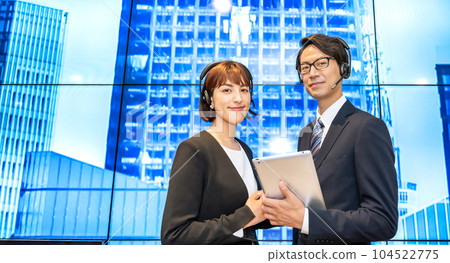 一位女商人和一位戴著耳機的商人在巨大的顯示屏前解釋 一位女商人和一位戴著耳機的商人在巨大的顯示屏前解釋 104522775