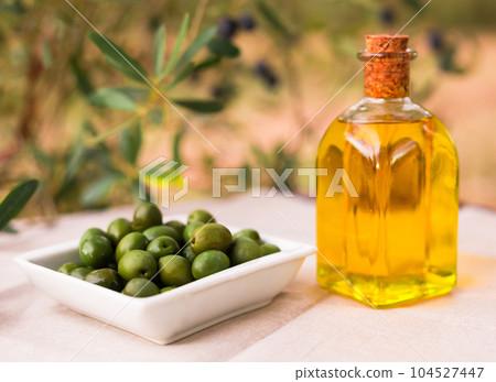 olive oil in a glass bottle and ripe black olives on the background of olive branches in the garden olive oil in a glass bottle and ripe black olives on the background of olive branches in the garden 104527447
