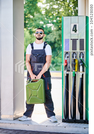 Professional worker holding canister in hands. Confident operator in sunglasses and overalls waiting for customers. Male worker standing on background of gas station with cistern in hands. Professional worker holding canister in hands. Confident operator in sunglasses and overalls waiting for customers. Male worker standing on background of gas station with cistern in hands. 104528080