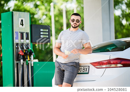 Young man waiting for petrol station operator to pay for fuel. Man leaning on his car and counting how much gasoline costs. Male in casual clothes holding money and looking straight into camera. Young man waiting for petrol station operator to pay for fuel. Man leaning on his car and counting how much gasoline costs. Male in casual clothes holding money and looking straight into camera. 104528081