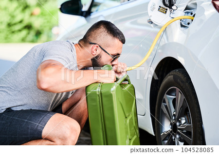 Weird man pumping gasoline from a gas tank into canister. Self-confident thief stealing fuel in broad daylight. Male adult trying to pump fuel from tank by hose. 104528085