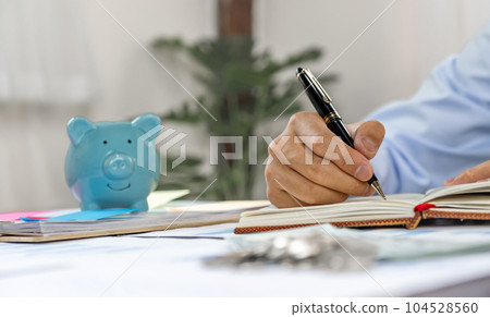 Businessman hand writing income and expenses to account with piggy bank and dollar notes, step up start-up businesses to success, Saving money for future plan and retirement fund concept 104528560