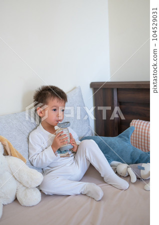 Toddler having his morning milk while sitting on the bed drinking from a bottle with a straw surrounded by his toy friends 104529031