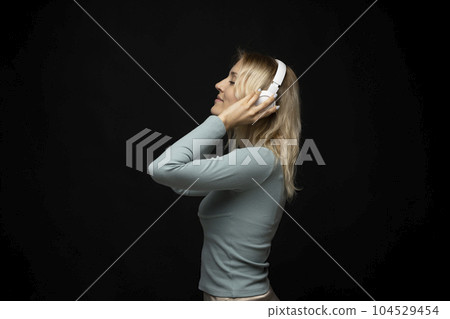 Profile portrait of charming blonde woman in massive white headphones listening to music on isolated black background. 104529454