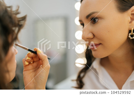 Closeup of female makeup artist applying cosmetic tonal foundation for face sculpting of pretty young woman in beauty studio. Creating creative image for photoshoot. Make-up artist corrects make-up. 104529592