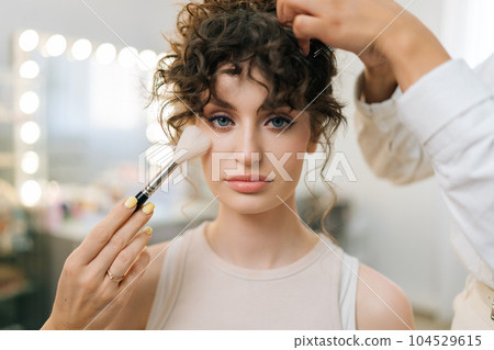 Closeup portrait of young elegance woman looking at camera with sensual expression while makeup artist and hairdresser final touch and corrections, of great make-up and hairstyle, bokeh lights. 104529615