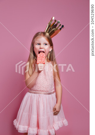Cute little kid girl in pink dress and a crown eating ice-cream on pink background 104529690