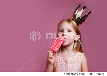 Cute little girl in a fancy dress and a crown eating ice-cream on pink background 104529691