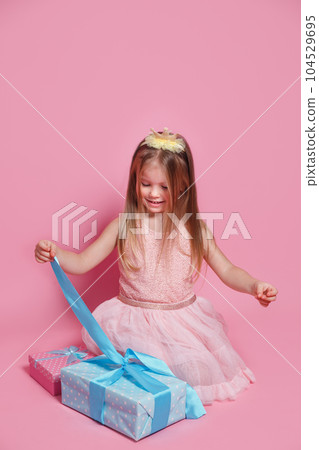 Cute excited kid girl opening present box sitting on pink studio background Cute excited kid girl opening present box sitting on pink studio background 104529695