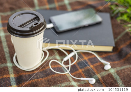 Cup of coffee, book and smartphone with headphones on blanket in park or garden Cup of coffee, book and smartphone with headphones on blanket in park or garden 104531119