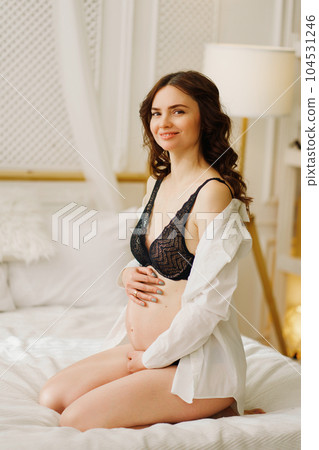 a slender pregnant woman in a black underwea and white shirt on the bed 104531246