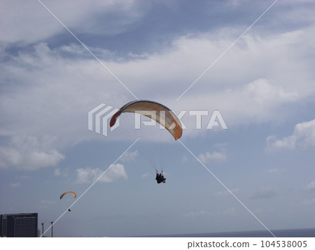 Two paragliders are flying high in the sky. Only the silhouette is visible. Against the background of a blue sky with clouds. Two paragliders are flying high in the sky. Only the silhouette is visible. Against the background of a blue sky with clouds. 104538005