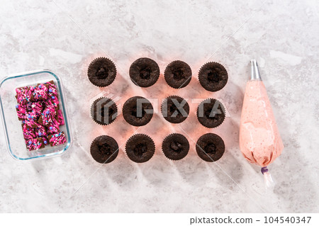 Flat lay. Piping strawberry buttercream frosting on top of chocolate cupcakes. 104540347