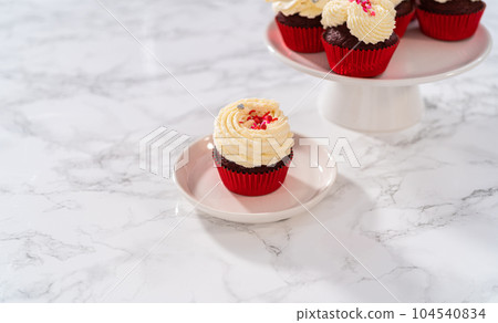 Freshly baked velvet cupcakes with white chocolate ganache frosting decorated with sprinkles. 104540834