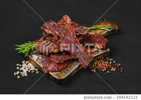 Dried beef jerky slices on wooden board with salt and pepper. Dried beef jerky slices on wooden board with salt and pepper. 104542115