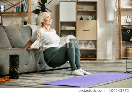 Smiling, beautiful, senior woman training at home in living room, doing exercises with couch. Strong fit body 104543773
