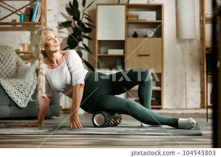 Slim, sportive, beautiful senior woman training at home, keeping body in tone with fitness massage roller. Slim, sportive, beautiful senior woman training at home, keeping body in tone with fitness massage roller. 104543803