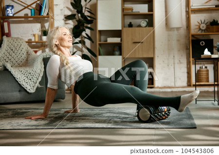 Slim, sportive, beautiful senior woman training at home, keeping body in tone with fitness massage roller. 104543806