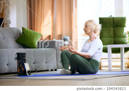 Beautiful, relaxedsenior woman in stylish sportswear sitting on fitness matt, doing yoga exercises at home in the morning Beautiful, relaxedsenior woman in stylish sportswear sitting on fitness matt, doing yoga exercises at home in the morning 104543821