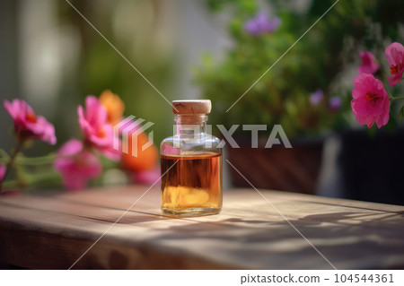 Fragrant Rose Geranium Essential Oil in small transparent glass Bottle on a wooden surface next to fresh Pink Geranium Flowers in full bloom, Garden background with Copy space. 104544361