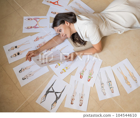 A woman lies on the floor and looks at sketches of swimwear and underwear.  104544610