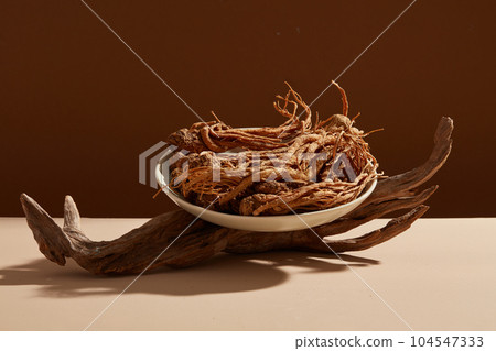 Front view of angelica sinensis roots on round white plate and dry twig decorated on dark background. Scene for medicine advertising, photography traditional medicine content Front view of angelica sinensis roots on round white plate and dry twig decorated on dark background. Scene for medicine advertising, photography traditional medicine content 104547333