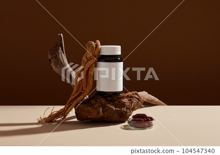 Minimalist scene for advertising and branding medicine product with herbal ingredient. Angelica sinensis roots with an empty bottle unbranded and capsules pills on brown background. Minimalist scene for advertising and branding medicine product with herbal ingredient. Angelica sinensis roots with an empty bottle unbranded and capsules pills on brown background. 104547340