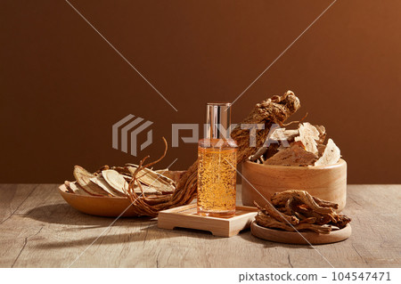 A serum glass bottle filled with yellow essence standing on a podium. Angelica root and Bai Zhu are contained in some wooden objects 104547471