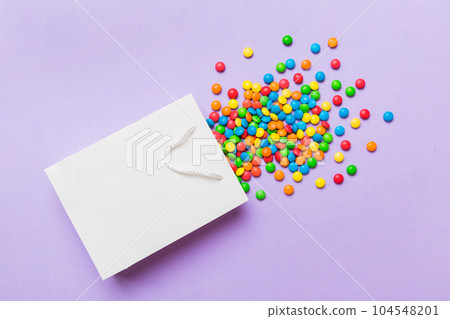 shopping paper gift bag in corner full of assorted traditional candies falling out on colored background with copy space. Happy Holidays sale concept shopping paper gift bag in corner full of assorted traditional candies falling out on colored background with copy space. Happy Holidays sale concept 104548201